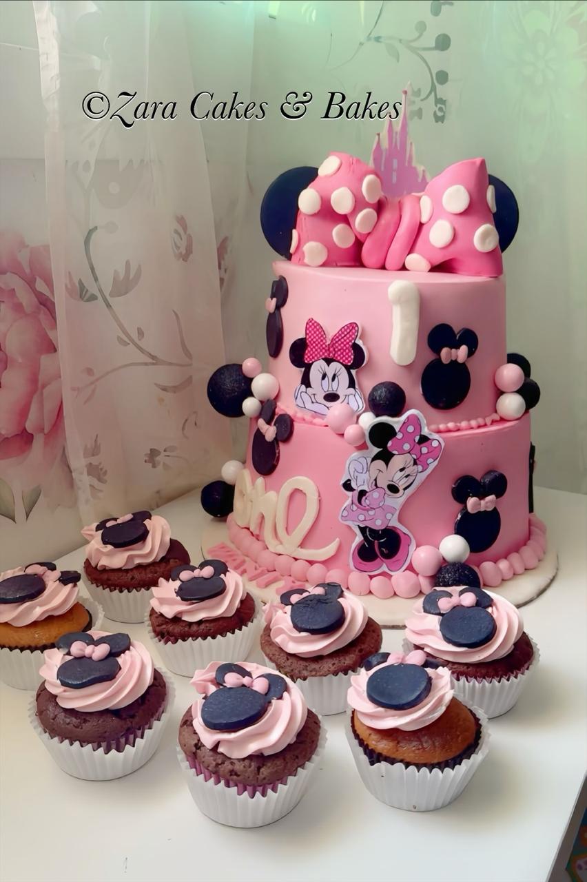 Two-tier pink Minnie Mouse first birthday cake with polka dot bow and matching themed cupcakes