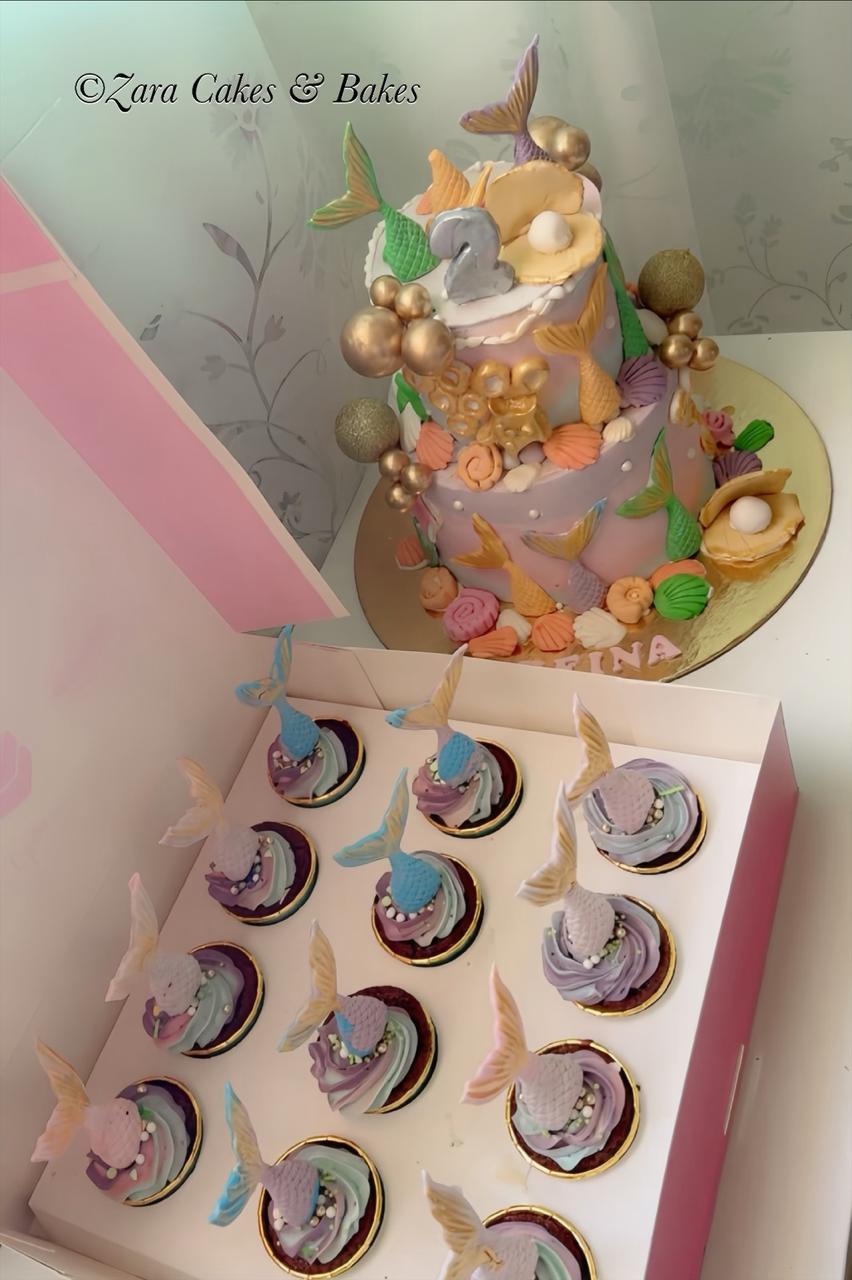Pastel purple mermaid birthday cake with gold shells, pearl decorations, and matching mermaid tail cupcakes in box