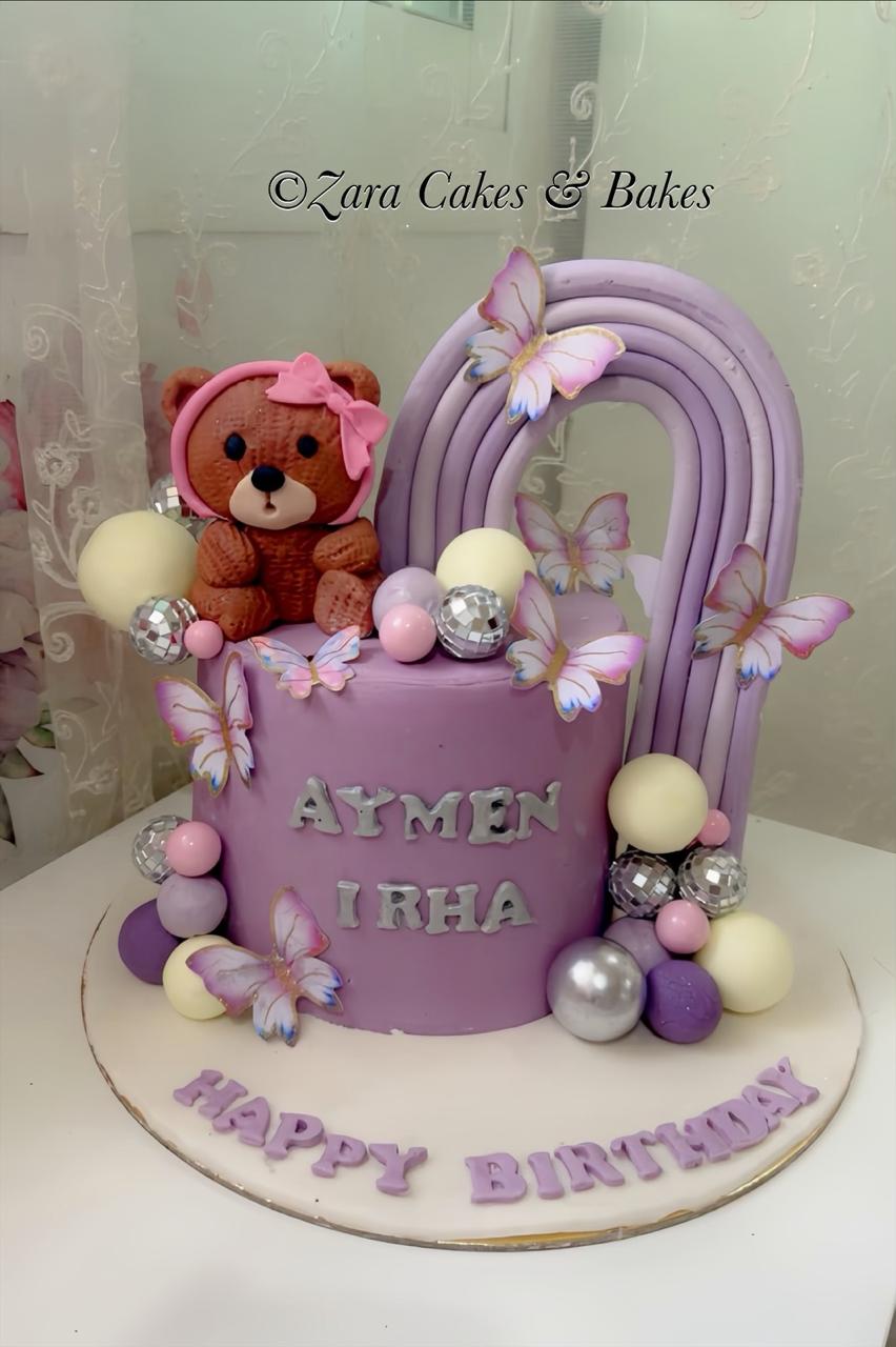 Lavender fondant birthday cake with teddy bear topper, rainbow arch, and butterfly decorations