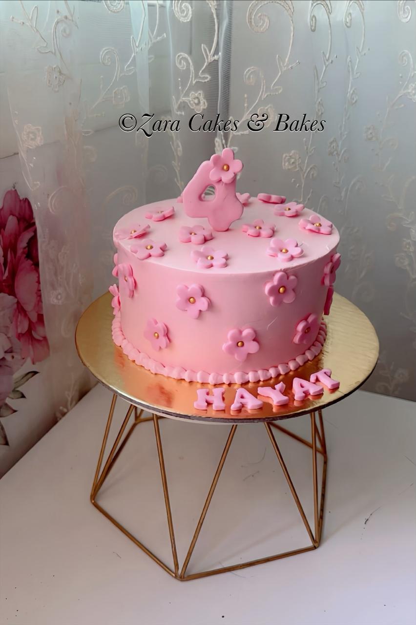 Pink fondant birthday cake with floral decorations, number 4 topper, and name Hayat on gold stand.