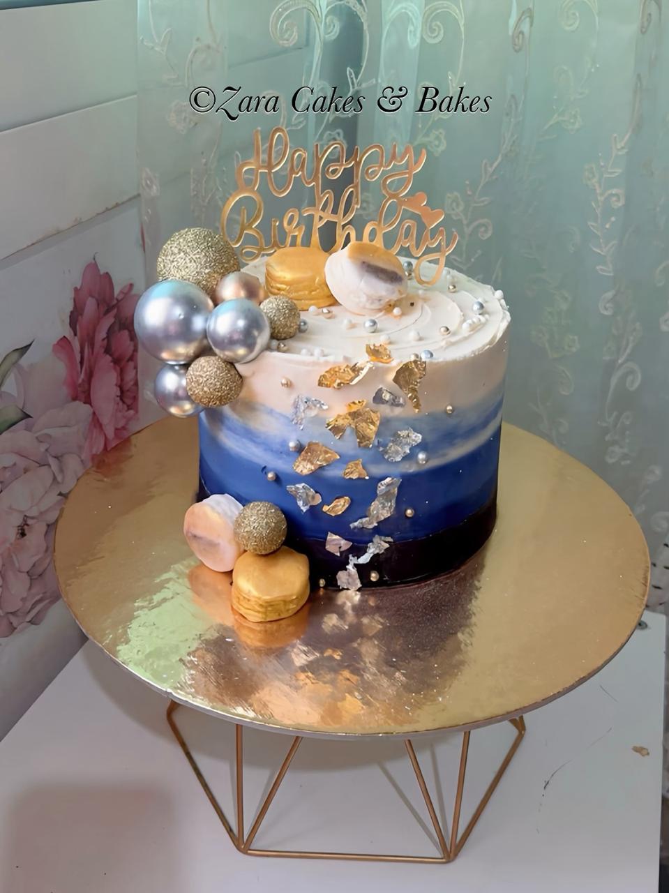 Blue ombre birthday cake with gold leaf flakes, macarons, and silver pearl decorations on a golden stand