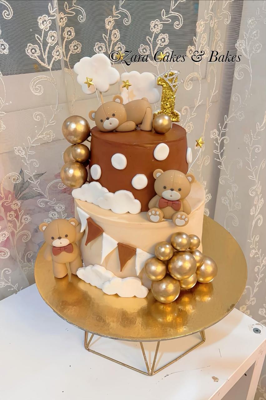 Two-tier teddy bear first birthday cake with chocolate and vanilla tiers, gold balloons, and cloud decorations