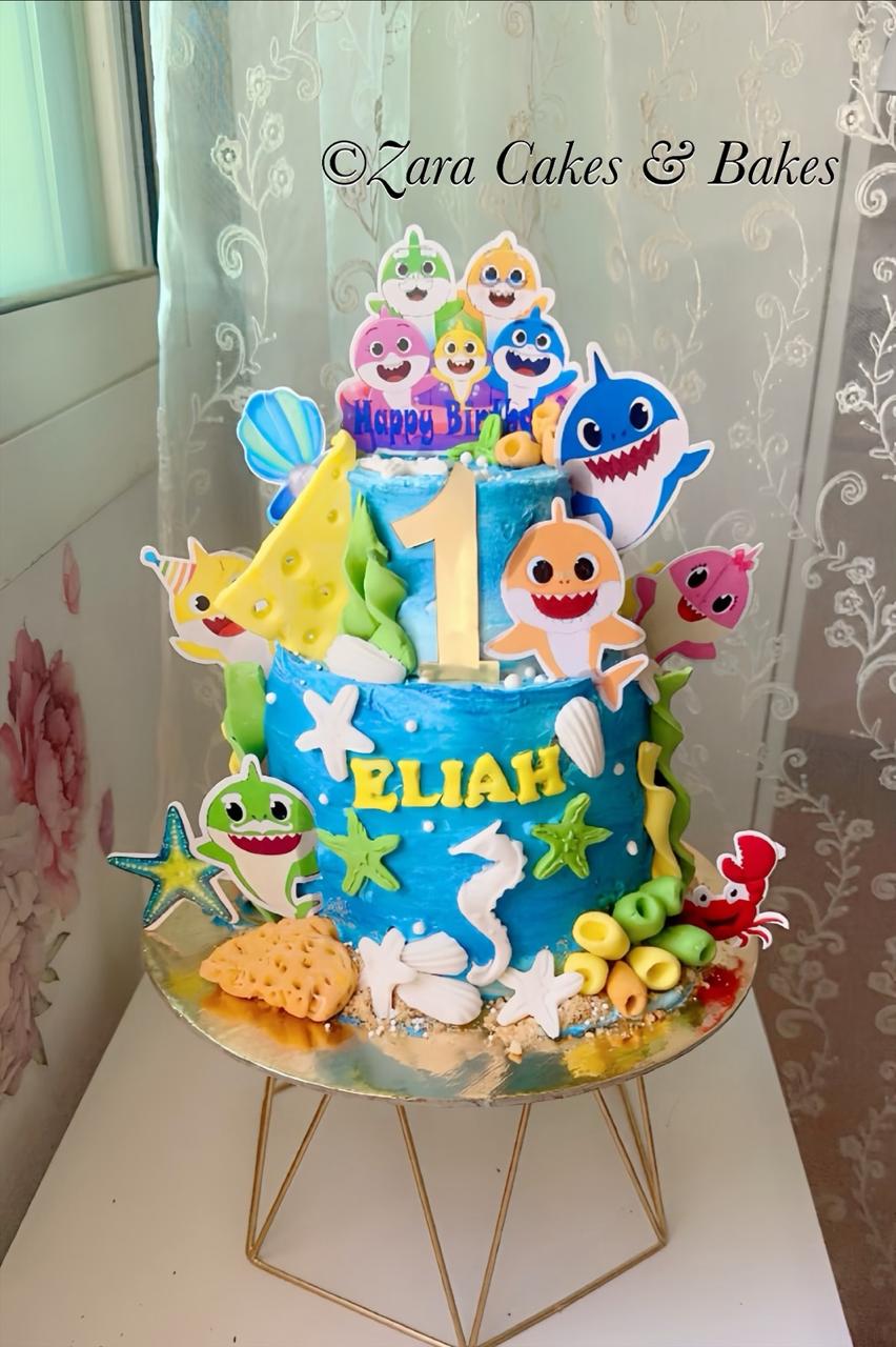Two-tier Baby Shark first birthday cake with blue buttercream, fondant sea animals, and gold number topper for Elijah.