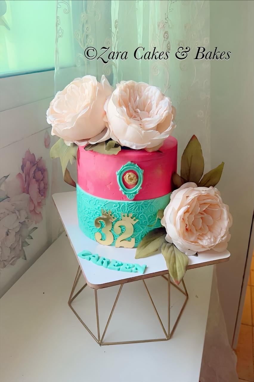 Pink and teal two-tier birthday cake with white sugar flowers, gold number 32, and vintage cameo decoration