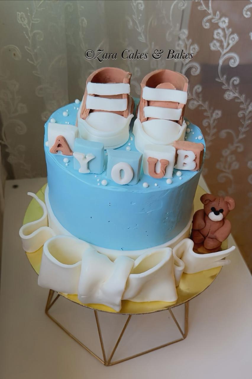 Blue baby shower cake with fondant booties, name blocks, teddy bear, and white ribbon bow decoration