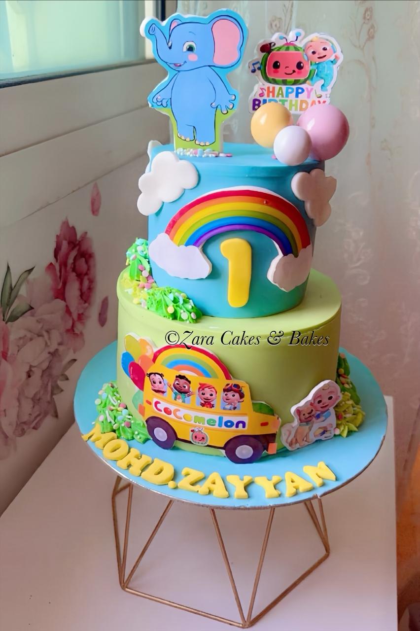 Colorful two-tier Cocomelon 1st birthday cake with rainbow, elephant topper, and yellow bus decoration